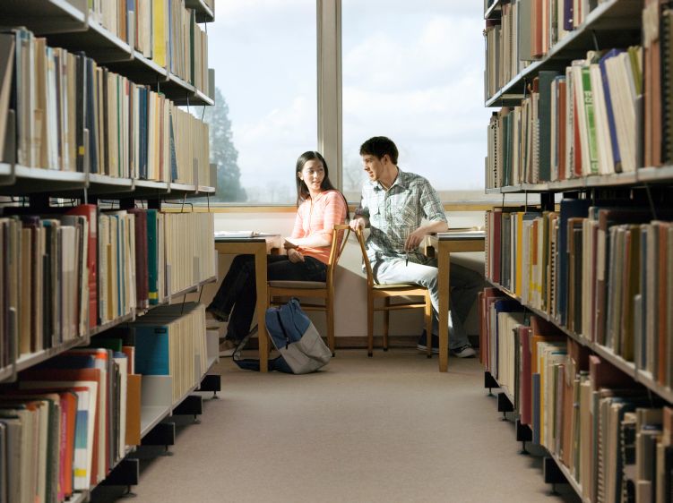 2 college students sitting next to the window in a library talking to each other
