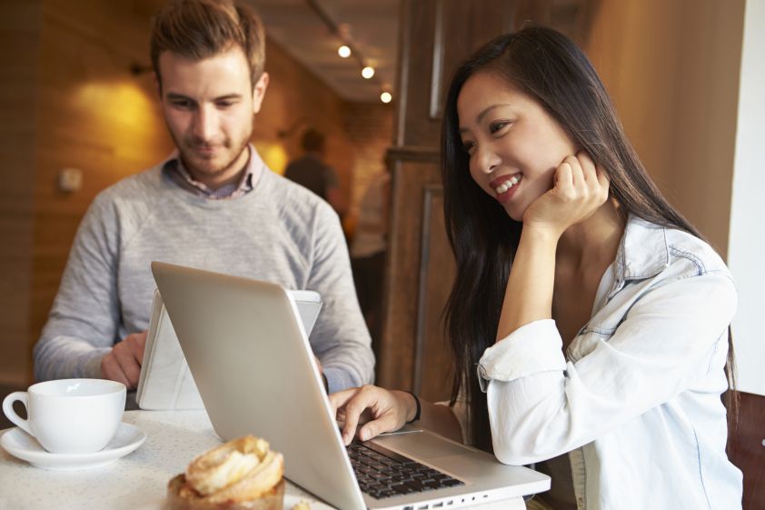 Smiling Oriental woman sitting in front of laptop smiling next to a white male on an ipad having coffee